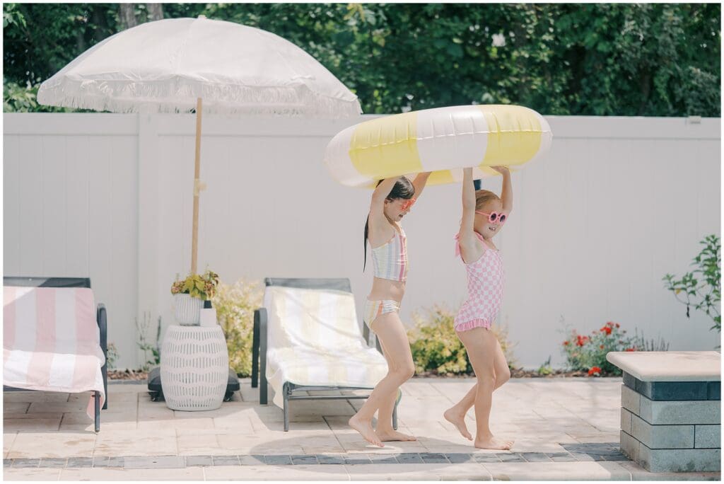 Two little girls carry a yellow and white pool float over their heads during a summer splash photo session with Jessica Jeremiah Photography